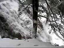A video taken in winter from the trail starts by looking up to the head of the waterfalls and then pans slowly down and to the right to follow the falling water cascading down a stone face. The thundering water flows freely, but the surrounding rocks and trees are covered in snow and ice.
