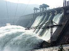 Image of a large water retaining structure that spills water like a waterfall, some power lines and a construction crane