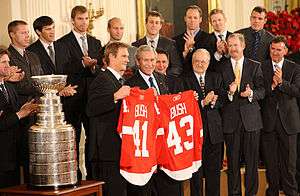 In the front row, Nicklas Lidstrom holds a Red Wings jersey with the last name "BUSH" and the number "41" while George W. Bush holds a Red Wings jersey with the last name "BUSH" and the number "43". To Lidstrom's right is the Stanley Cup. 2nd row, left to right: Mike Babcock, Ken Holland, Jim Devellano, Jim Nill, unknown man. Third row, left to right: Kris Draper, Pavel Datsyuk, Mikael Samuelsson, Brian Rafalski, Derek Meech, Kirk Maltby, Chris Osgood, Aaron Downey.