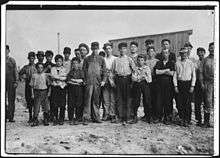 20-25 males facing camera in a row, mostly children. Ages vary. They are in work clothes. Some are dirty. Some wear caps. They stand on dirt with a wooden building behind them.