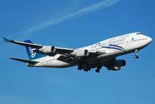 An Air New Zealand 747-400 with its landing gear down and flaps down. The aircraft is mostly white and blue aircraft and in-flight against a blue sky. On each of the two wings are two engines.