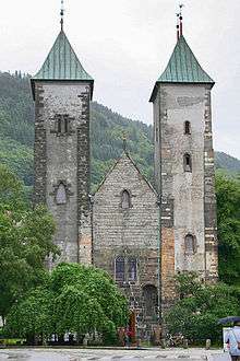 Stone Romanesque church with two towers and a lower, pitched roof entrance hall between them. The towers have slightly curved bronze roofs.