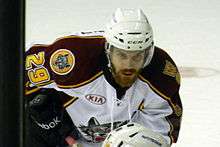 A Caucasian ice hockey player bent over at the waist. He wears a white helmet and has a large beard.