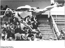 A man in an athletic uniform is diving forward, face-down over a high jump bar. His right leg is extended straight, parallel to the bar, while his left is bent at the knee and is just touching the bar as he leaps over it.
