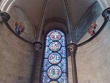 A semi-circular area with stone walls, a domed ceiling and a tall stained glass window with prominent circular decorations.