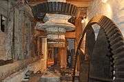 on the floor below the stones is the drive from the wheel.  We see the same vertical shaft that goes up through the floor, topped here with a black iron bevel gear.  Another bevel gear on the right, currently disengaged, is driven by the waterwheel. The room is cramped and whitewashed.