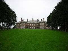 A large mansion house is framed by rows of trees on either side and an expanse of well-kept green lawn in front.