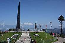 An obelisk on an elevated circular platform, with a few people standing around. Kangchenjunga is visible in the background.