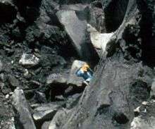 A man climbing a steep cliff face dwarfed by massive boulders around him.