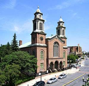 A brick building with two tall towers topped with copper, seen from its right, across a city street, at some height. Other buildings are visible on the right of the image.