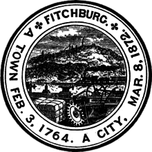 A black and white circular seal with a group of gears and mechanical parts in the foreground and a hill with a rock on the top in the background.
