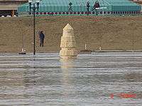 Water from a river completely surrounds a stone obelisk. In the background are several people with news cameras. A building with a green roof is seen behind them. At the bottom right of the image is the camera's time: "6 7:59 am".