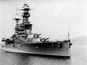 A three-quarter view of a heavily-armoured battleship at anchor. There are two main turrets visible before the bridge, each housing a pair of 15-inch guns. 6-inch guns are housed in a row of individual sideways-facing sponsons. The flank of the ship has a conspicuous bulge at the waterline