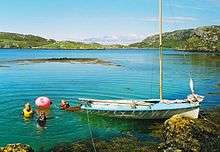 A small blue sailing dinghy lies in an aquamarine body of water next to a rocky shore underneath light blue skies. Three people are swimming off the stern of the boat next to a red buoy.