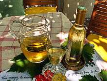 Colour photo illustrating well the song "Ah, the little white wine ...". A carafe of white wine with a full glass and a corked bottle is placed on vine leaves, themselves on a white tablecloth with red tiles. The bottle covered with mist gives a feeling of freshness and the play of light and shadow suggests that the scene is set under a Pergola.