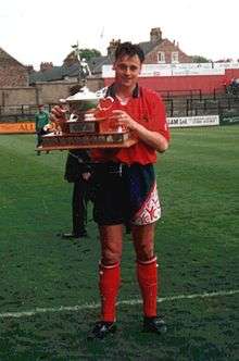 A man standing on a grass football pitch, wearing a red shirt, black shorts and red socks, and holding a trophy.