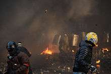 Three helmeted protestors throwing pavement bricks at riot-police line under concealment of smoke from burning tires