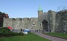 Quebec's restored city wall is gray stone about 20 feet (6.5 meters) high. The St. John's gate has a modern road going through it, and has a copper-roofed turret on the left bastion. A paved path goes through a grassy area below the wall.