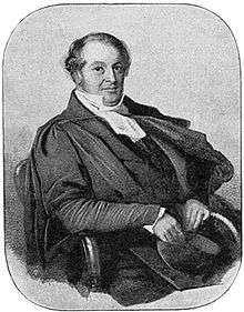 A balding middle-aged man in a dark suit and academic gown, with a high collared white shirt and academic bands around his neck. He is sitting in a chair, holding a mortar board