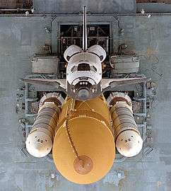 An overhead view of a spaceplane, coloured white on its topside and black on its underside, attached to a large orange tank, to which two slender white rockets are also attached. A gray platform supporting this stack serves as the background.