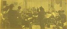 A somewhat indistinct photo on yellow paper of a crowded meeting room, showing student protesters with signs. In the front, several well-dressed individuals appear to be gathering papers and departing.