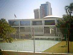 Photograph of the Supreme Court Building, a bright white building of two stories with an open space below, overtopped by a shallow dome, but obscured by a foreground fence