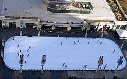 An ice skating rink with a few skaters and a large metallic sculpture in the background