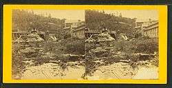 Old black and white photo of a waterfall, flanked on both sides with slightly dilapidated-looking buildings.