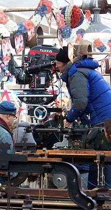 A man wrapped in a blue jacket and black hat looks down the scope of a large film camera. There is red, white, and blue bunting hanging overhead.