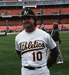Man standing on a baseball field looking slightly to the left. He is wearing a gray Oakland Athletics uniform with green sleeves and a gree and yellow Athletics hat.