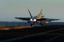 Photograph from behind a twin-engined jet fighter. The aircraft's wheels are on the surface, but the engines are still active, and a hook on the underside of the aircraft is in contact with the surface and trailing sparks
