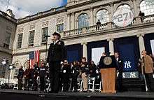 A woman in an American Navy uniform stands before a microphone with several people behind her on stage. The stage is in front of a building with a sign that reads "#27!".