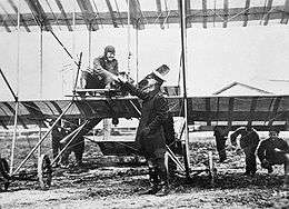Pilot seated in open-framed biplane, shaking hands with a man in military uniform who wears a peaked cap