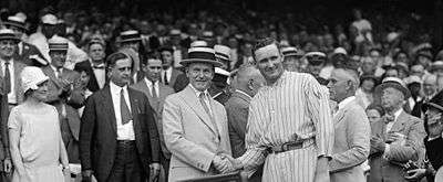 Two men stand in front of a crowd shaking hands. The man on the left of the photo is wearing a tan suit and hat and the man on the right is wearing a light-colored pinstriped baseball uniform.
