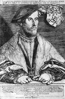 A  middle aged man, Wilhelm, Duke of Jülich-Cleves-Berg, is sitting at a table. He is  dressed in a soft cap that falls to the side of his head. He is wearing  fur-trimmed  robes, and nestled in the edges of the robes is a chain, with a cross  at its nadir. His hands are folded on the table before him. He is  wearing several rings, and one hand holds a pair of gloves. The family  crest hangs on the chair behind him.