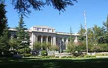 A large white stone governmental building, with pillars in front. Parts of the facade are obscured by evergreen trees.