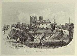 Trains passing beneath a long wall to enter a station, near a large cathedral